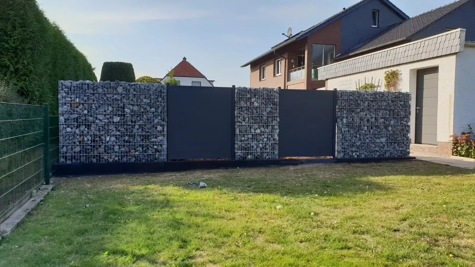 Sichtschutzzaun mit befüllbaren Drahtkörben und dunkelgrauen Kunststoff-Elementen.